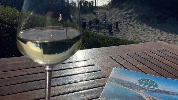 Um copo de vinho branco sobre uma mesa de madeira, com vista para a praia ao fundo.
