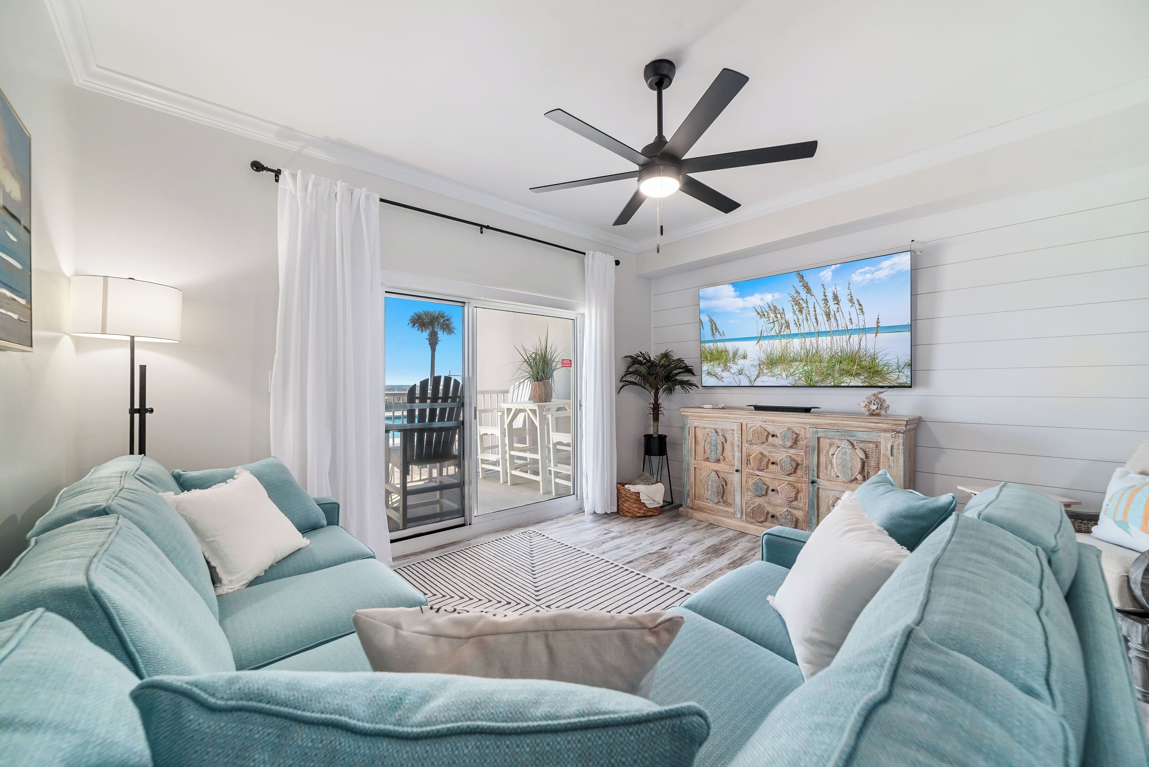 A living room with a teal sofa, a ceiling fan, a TV, and a view of the outdoors.