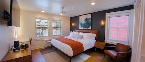 A modern bedroom with a large bed, a desk, a chair, and a window with a view of the street.