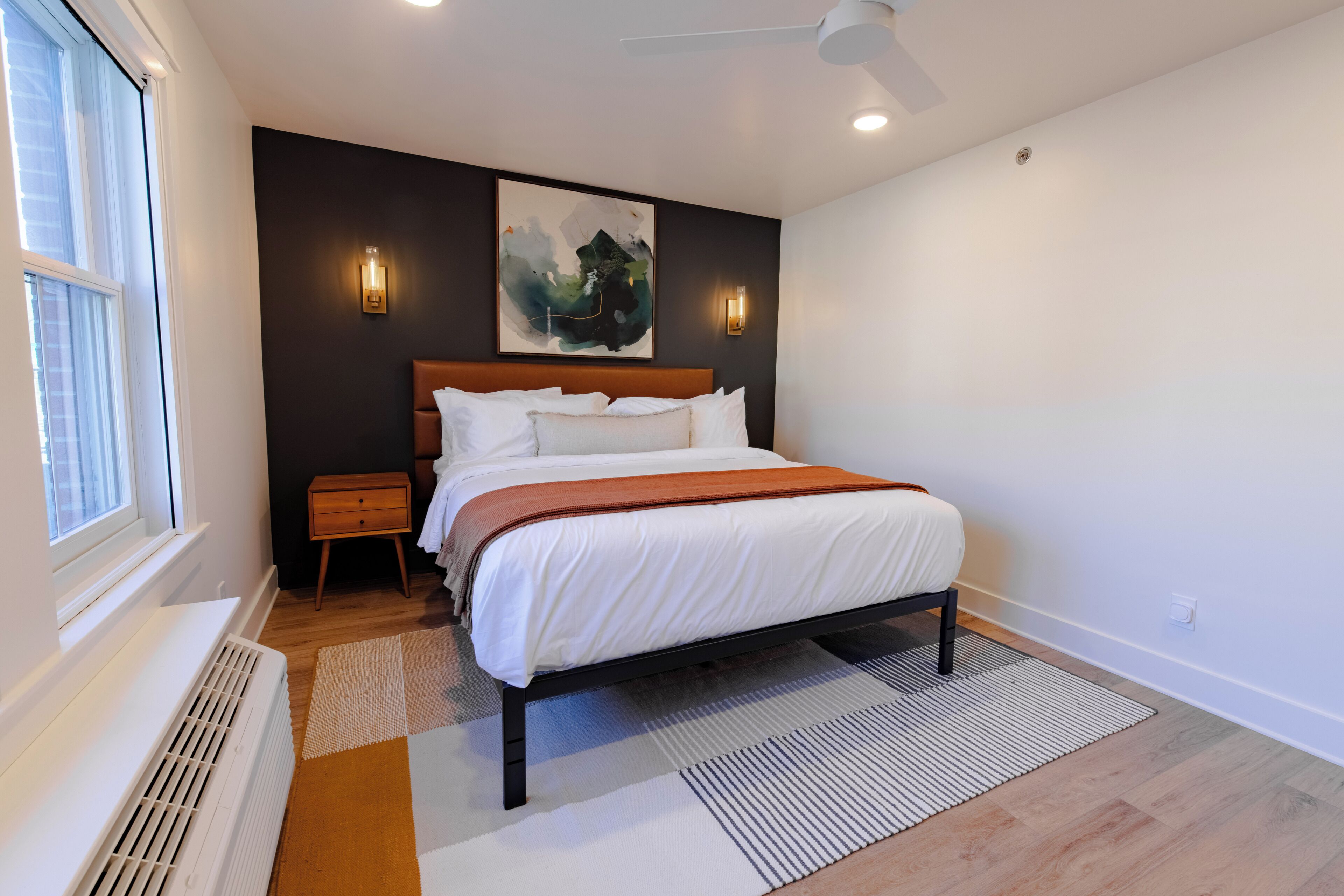 A modern bedroom with a large bed, a nightstand, a wall-mounted artwork, and a window with blinds.