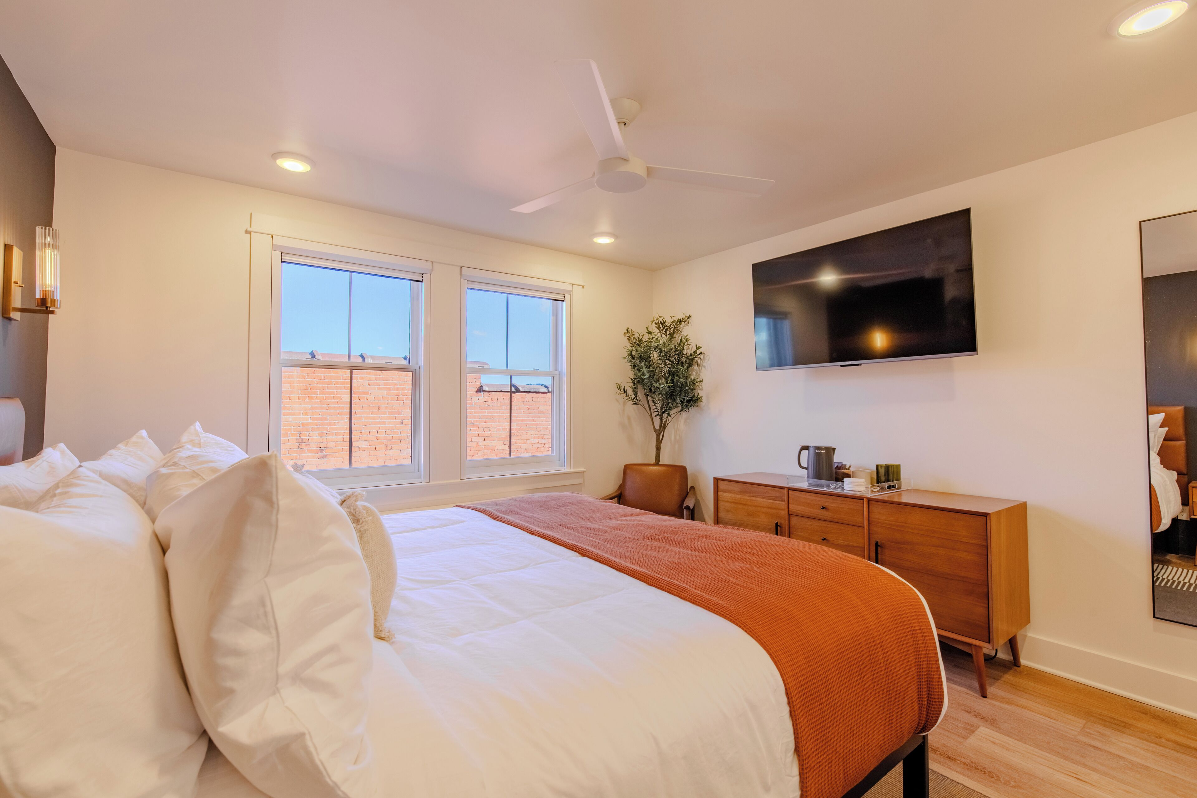 A modern bedroom with a large bed, a wooden dresser, a flat-screen TV, and a window with a view of a brick building.