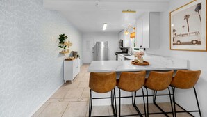 Une cuisine moderne avec des meubles blancs, un réfrigérateur en acier inoxydable et un îlot en marbre agrémenté de quatre tabourets de bar marron.