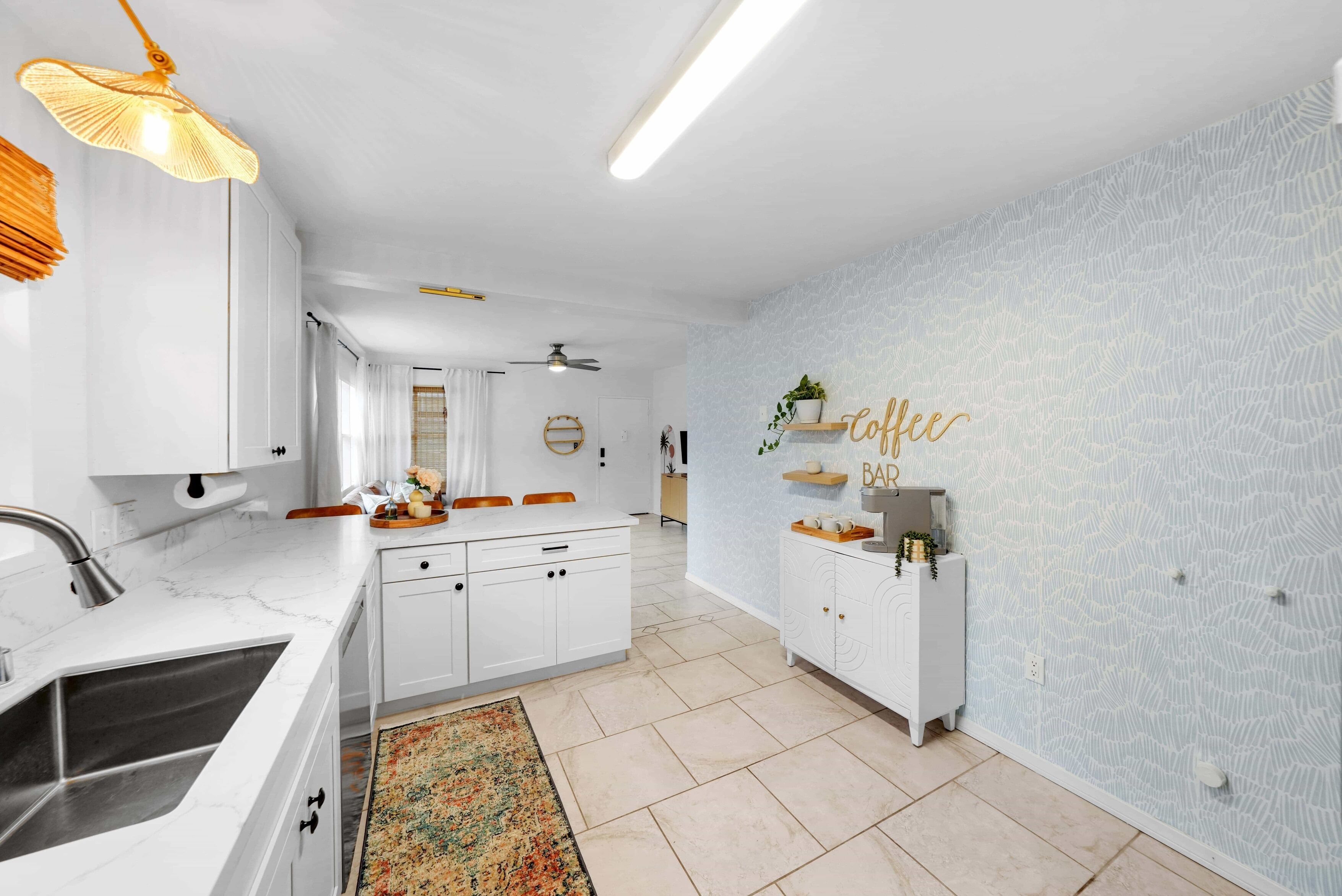A modern kitchen with white cabinets, a marble countertop, and a ceiling fan. There is a rug on the floor and a wall with a coffee bar sign.