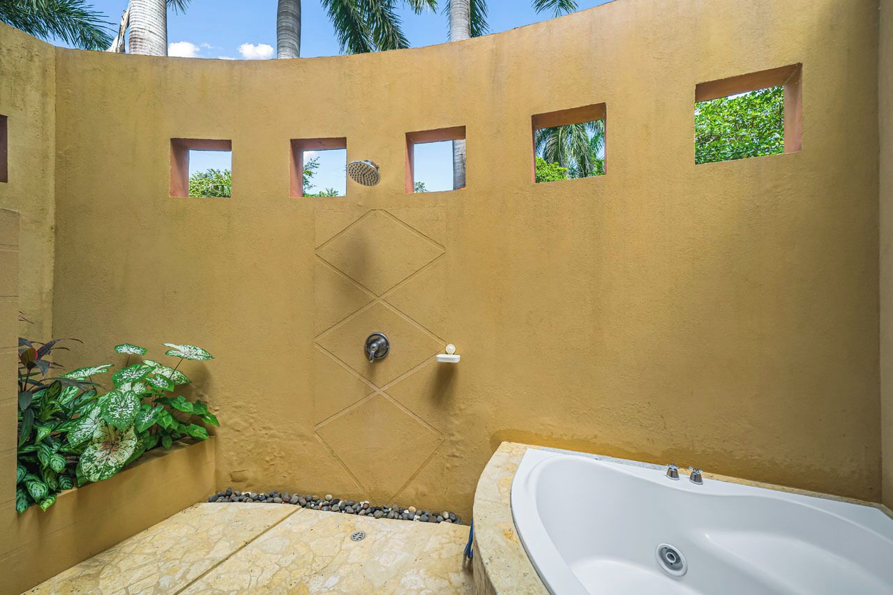 Un bagno con una vasca, una doccia con finestra a forma di diamante e una piccola finestra con vista su palme.