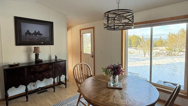 A dining area with a round wooden table, chairs, a hanging light fixture, a television on a cabinet, and a window with a view of trees and a snowy landscape. - Charming 3-bedroom house with AC, WiFi (Columbia Falls)