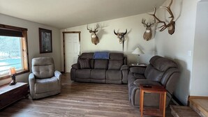 A spacious living room with a large sectional sofa, a recliner, and a wooden coffee table. There are mounted deer heads on the wall, and a window provides natural light. - Charming 3-bedroom house with AC, WiFi (Columbia Falls)