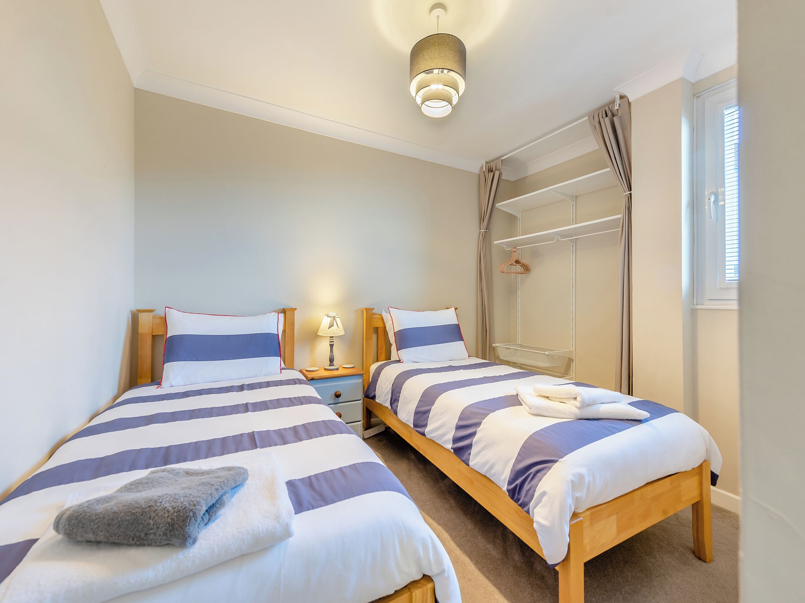 A bedroom with two beds, a bedside table with a lamp, a hanging shower curtain, and a window with blinds.