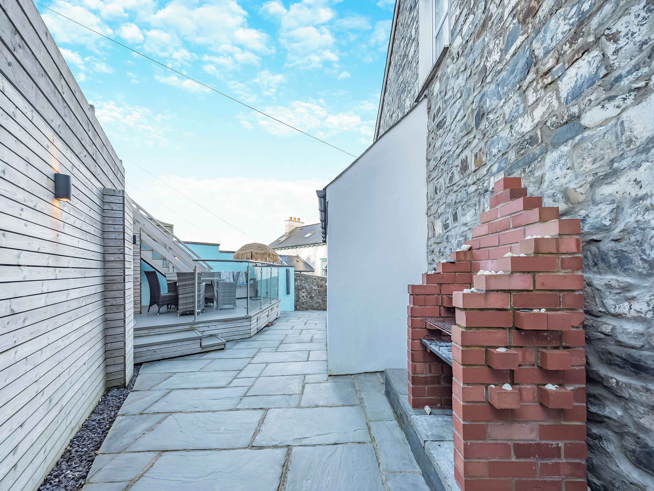 A narrow stone pathway leading to a patio area with a brick oven and outdoor furniture.