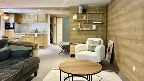 A modern living room with a sofa, a round coffee table, and a white armchair. There is a kitchen in the background with wooden cabinets and a built-in oven.