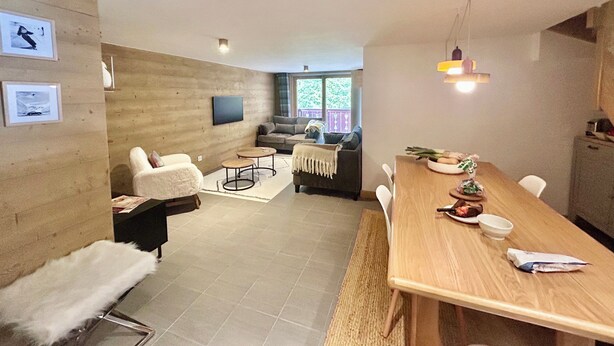 A modern living room with a sofa, coffee table, and dining table. There are framed pictures on the wall and a window with a view of greenery outside.