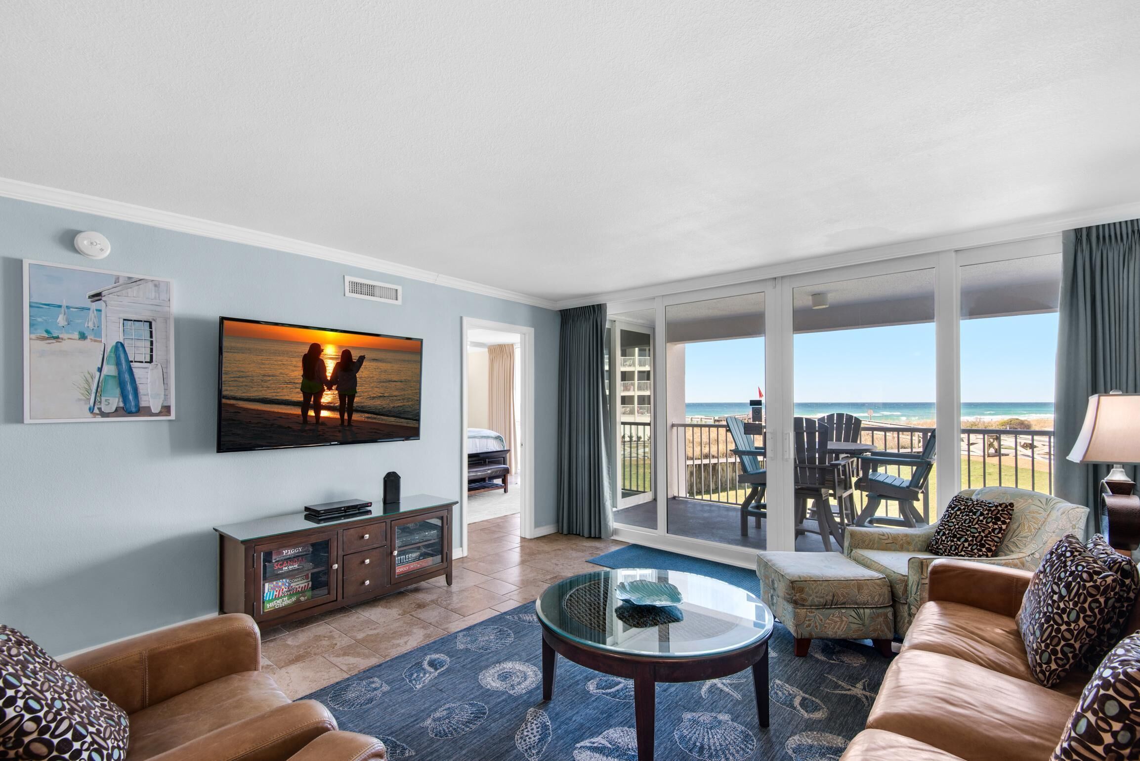 A living room with a large TV, comfortable seating, a glass coffee table, and a view of the beach through large sliding doors.