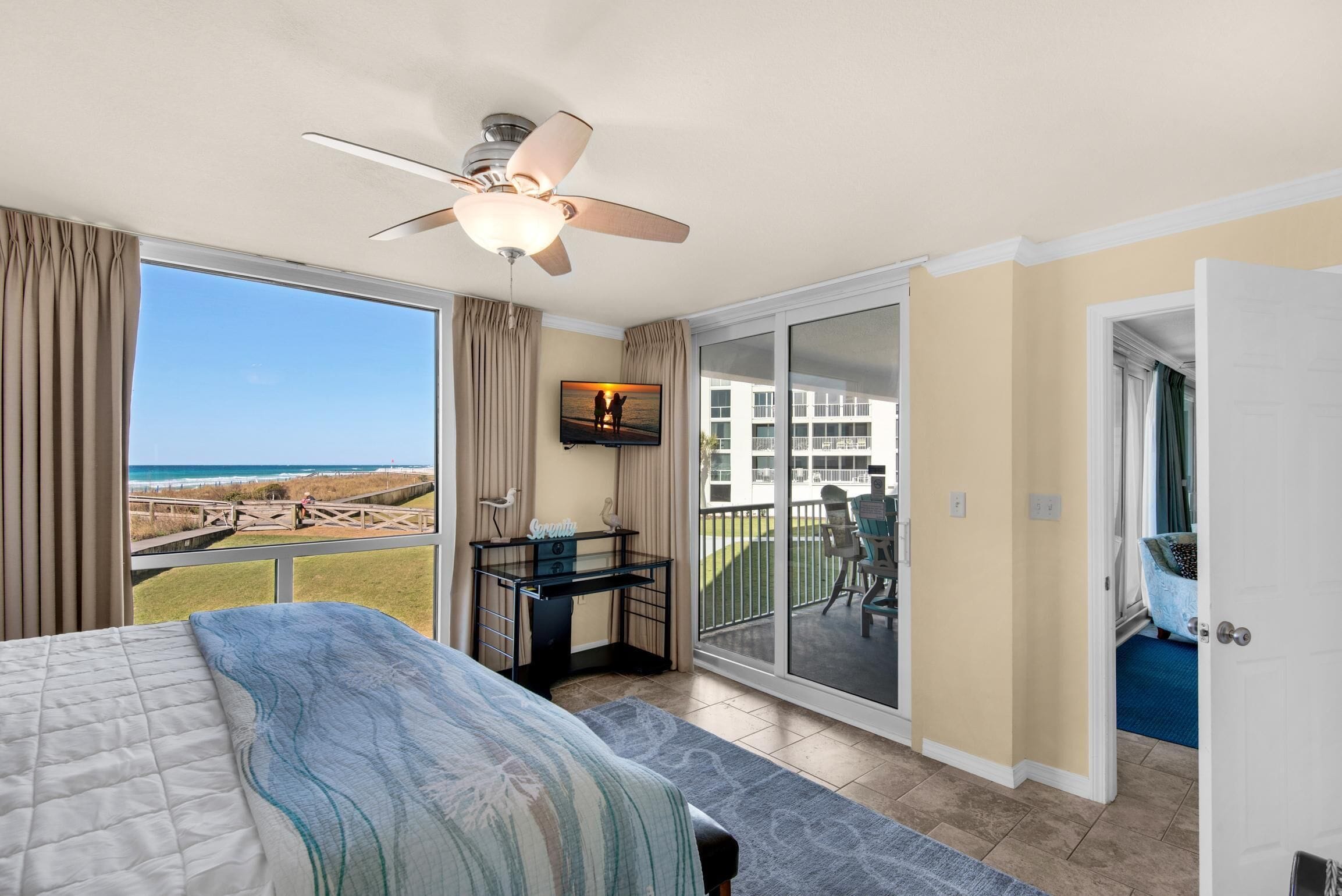 A bedroom with a bed, a desk, a ceiling fan, and a balcony with a view of the beach.