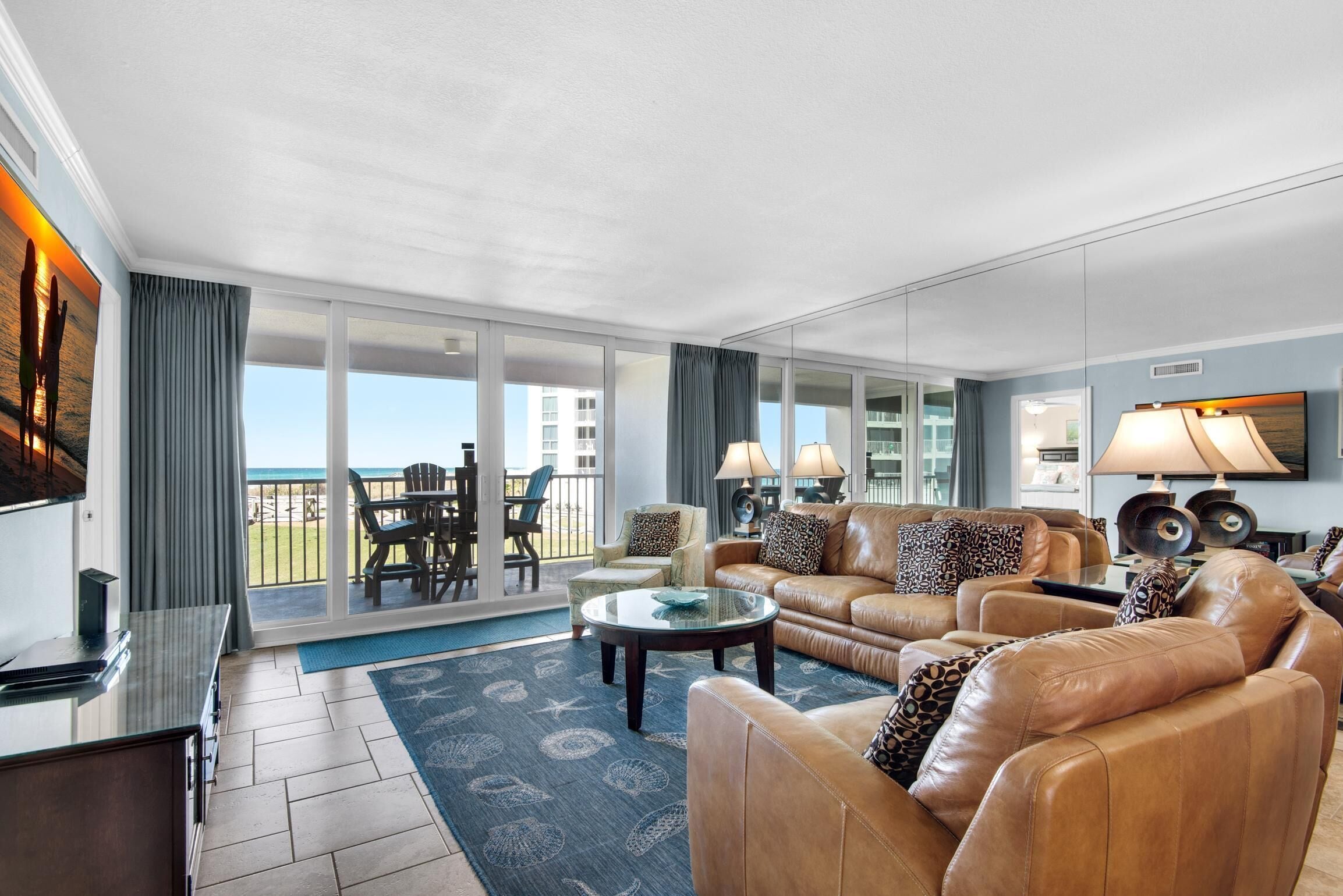 A spacious living room with a large sofa, a coffee table, and a TV. There is a balcony with a view of the ocean.
