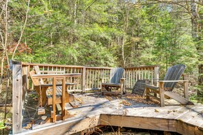 Puinen terassi, jossa on Adirondack-tyylisiä puutarhatuoleja ja pöytä, ja kaiken ympärillä on metsää.