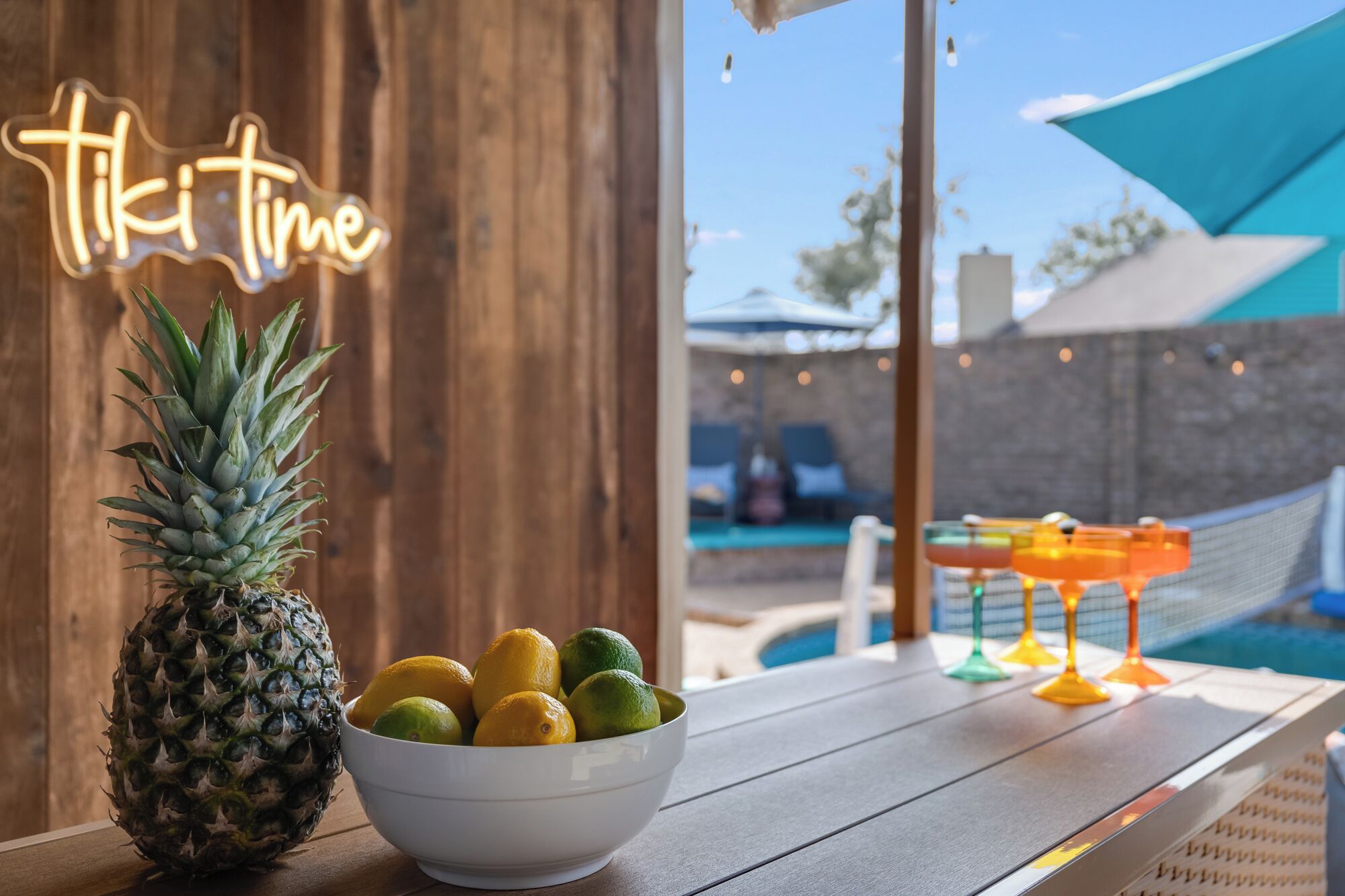 Una mesa de madera con un bol de fruta y una piña, un letrero de neón que dice "tiki time", y una piscina al fondo.