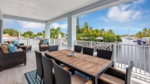 A covered outdoor area with a wooden table and chairs, a white railing, and a view of a marina with boats and palm trees. - Large Waterfront Escape, Canal Views (Tavernier)