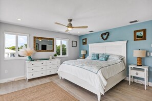 A bedroom with a bed, a dresser, a nightstand, a ceiling fan, and a window with a view.