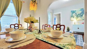 A dining room with a wooden table set for four, surrounded by chairs with heart-shaped backs. A vase with yellow flowers is on the table, and there is a painting on the wall.