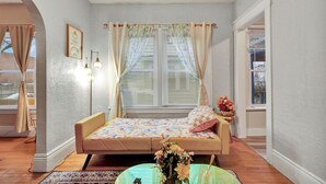 A bedroom with a bed, a window with curtains, a wall-mounted light fixture, and a small table with a vase of flowers.