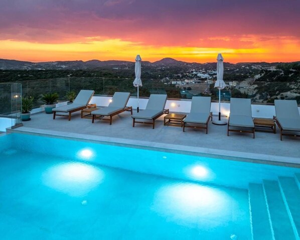 A poolside area with lounge chairs and umbrellas, overlooking a scenic landscape during sunset.