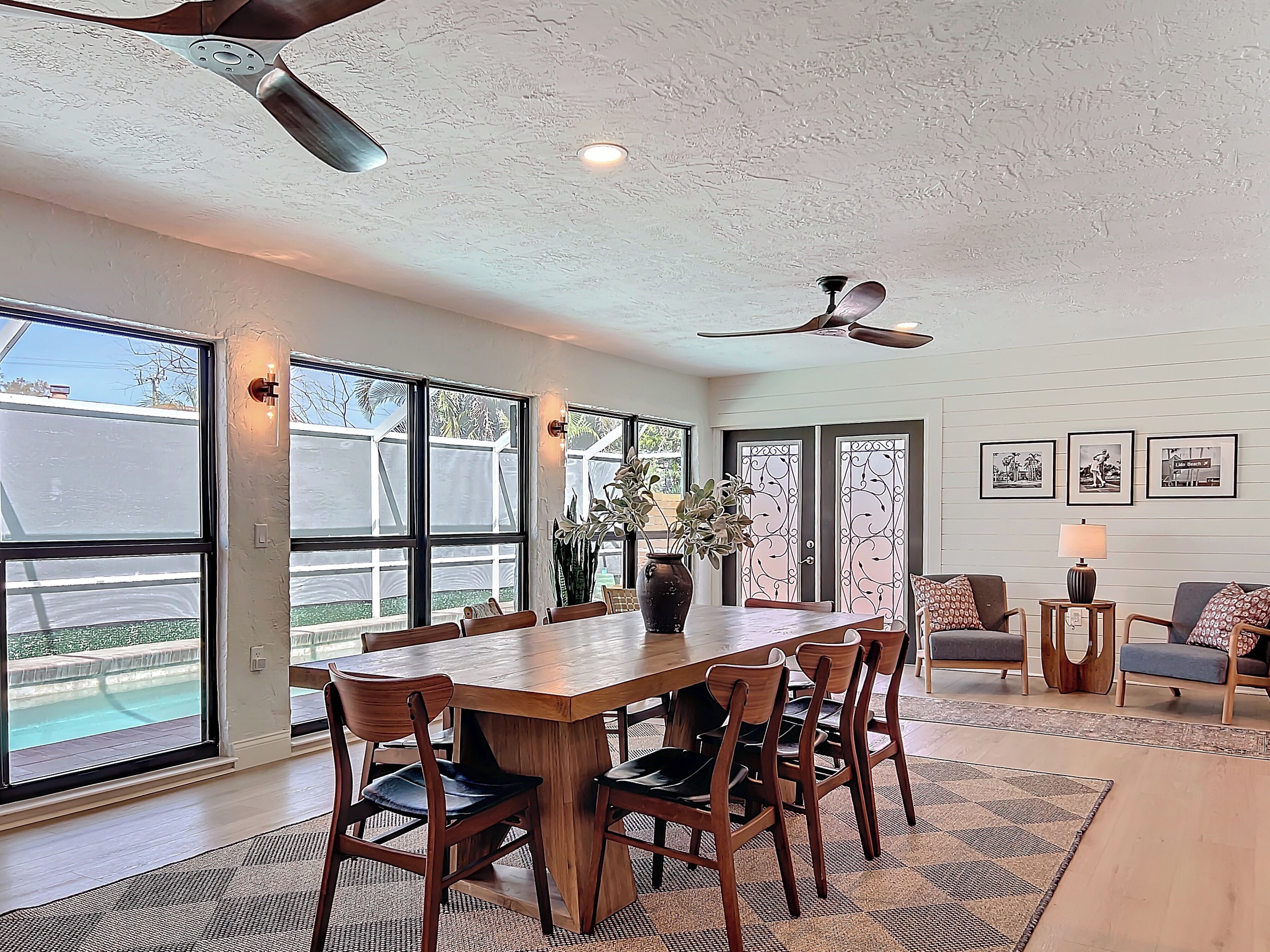 Un vaste espace à manger avec une grande table et des chaises en bois, de grandes fenêtres donnant sur une piscine, un ventilateur de plafond et une petite table d’appoint avec une lampe.