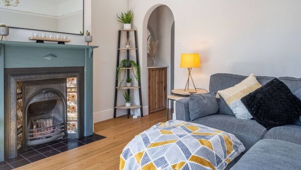 A living room with a fireplace, a sofa, a lamp, and a plant shelf. - Swift Host | Central | 3 Bedrooms | Parking (Norwich)