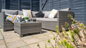 An outdoor seating area with wicker furniture, including a sofa and two side tables, set against a dark wooden backdrop. - Swift Host | Central | 3 Bedrooms | Parking (Norwich)