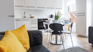 A modern kitchen with white cabinets, a dining area with a round table and chairs, and a living room with a black sofa and yellow pillow. - Swift Host | Park Lane | 2 Bed | Great Location (Norwich)