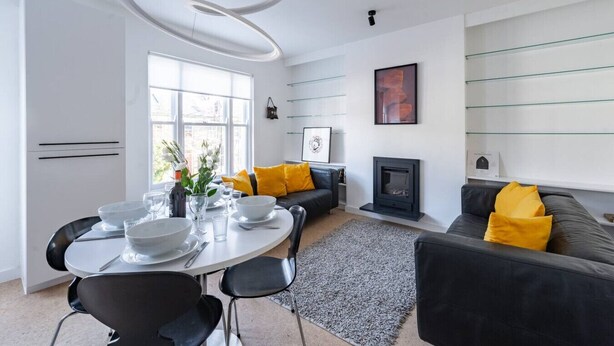 A modern living room with a fireplace, a dining area with a round table, and a sofa with yellow cushions. - Swift Host | Park Lane | 2 Bed | Great Location (Norwich)