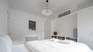 A minimalist bedroom with a large bed, a radiator, a bookshelf, and a pendant light. - Swift Host | Park Lane | 2 Bed | Great Location (Norwich)