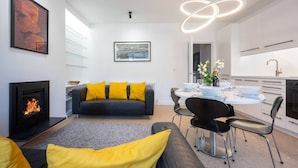 A modern living room with a fireplace, a sofa with yellow cushions, a dining area with a round table and chairs, and a kitchen with white cabinets and a pendant light. - Swift Host | Park Lane | 2 Bed | Great Location (Norwich)