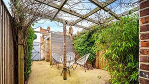 A covered patio area with two white wire mesh chairs, a wooden pergola, and dense foliage. - Swift Host | Stylish City Terrace | Parking (Norwich)