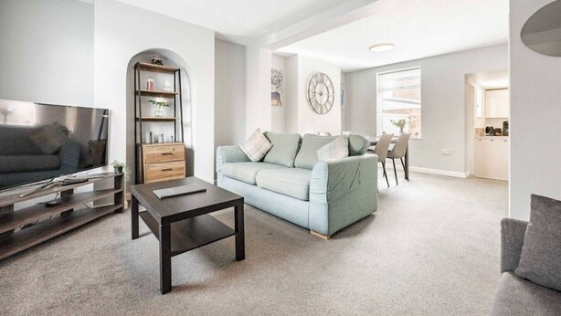 A modern living room with a flat-screen TV, a wooden entertainment unit, a light blue sofa, and a dining area with chairs and a table. - Swift Host | 2 beds | Central Location | Cowgate (Norwich)