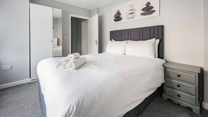 A neatly made bed with white linens and rolled towels, a grey headboard, and a bedside table with drawers. - Swift Host | 2 beds | Central Location | Cowgate (Norwich)
