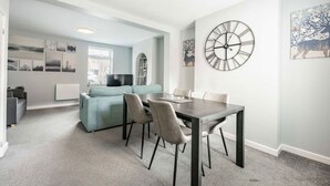 A modern living room with a dining area, a large wall clock, and artwork on the walls. - Swift Host | 2 beds | Central Location | Cowgate (Norwich)