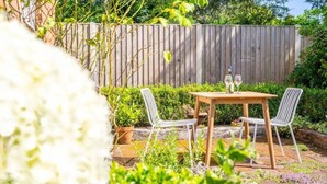 A patio with a wooden table and two chairs, surrounded by greenery and a wooden fence.