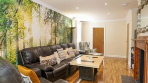 Un salon avec un canapé en cuir, une table basse et un fauteuil à motifs. Un grand décor mural représentant une forêt orne le mur.