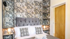 Un lit bien fait, avec une tête de lit grise, des oreillers blancs et une serviette pliée posée sur le lit. On peut voir deux tables de chevet avec des lampes, et une porte en bois à l’arrière-plan.