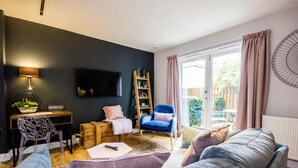 Ein modernes Wohnzimmer mit dunkler Wand, einem Fernseher, einem Holztisch, einem blauen Sessel, einem grauen Sofa und einem großen Fenster mit Vorhängen.