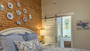 A bedroom with a brick wall, a bed with floral bedding, a white door, and a stained glass window. - Charming 2 BR/2 bath in Historic District - Rent 1 Suite or Both/8-16 people! (Troy)