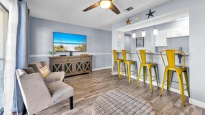 A living room with a sofa, a dining area with yellow chairs, a ceiling fan, and a kitchen area with white cabinets. - Endless Summer D5 (Panama City Beach)