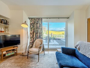 A living room with a TV, a sofa, a chair, and a view of a vineyard through a sliding glass door. - Newbigging Farm Cottage (Auchterarder)