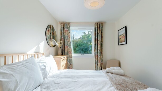 Une chambre à coucher avec un lit, une table de chevet, une lampe, une fenêtre avec des rideaux et un tableau encadré au mur.