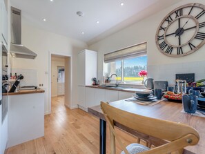 A modern kitchen with a large wall clock, wooden flooring, and a view of the outdoors. - Newbigging Farm Cottage (Auchterarder)