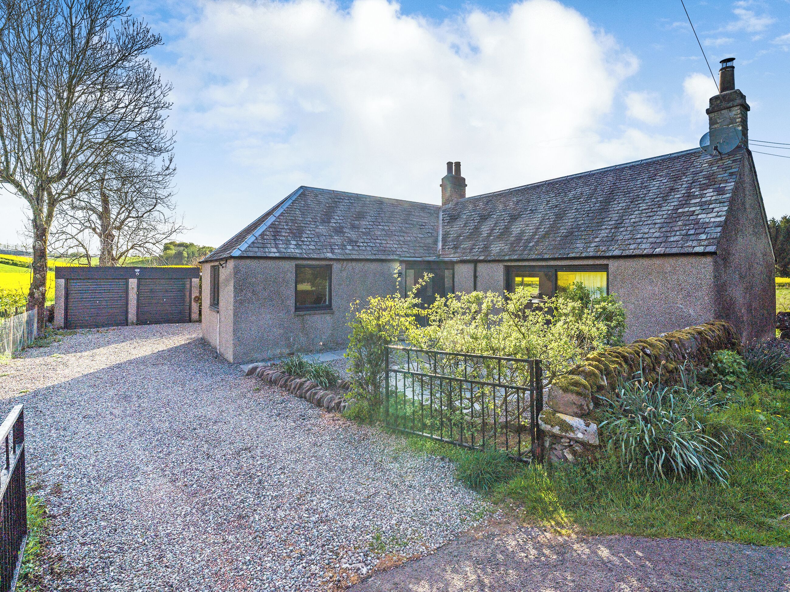 A stone house with a gravel driveway, a metal fence, and a garden with flowering plants.