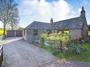 A stone house with a gravel driveway, a metal fence, and a garden with flowering plants. - Newbigging Farm Cottage (Auchterarder)