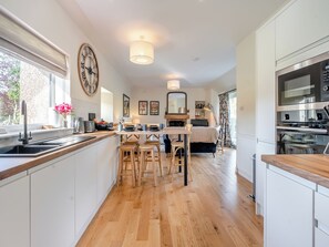 A modern kitchen with wooden flooring, a dining area with wooden chairs, a large clock, and a built-in oven. - Newbigging Farm Cottage (Auchterarder)