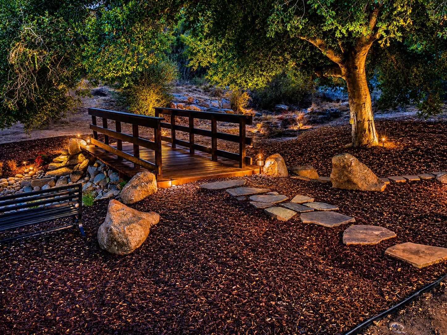 A wooden bridge over a gravel path with a stone border, surrounded by trees and rocks.