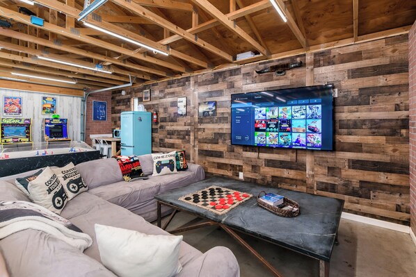 Ein Zimmer mit Holzbalkendecke, einem großen Flachbildfernseher an der Wand, einer Couch mit Gaming-Kissen und einem Schachbrett auf einem Tisch.