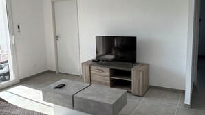 A modern living room with a flat-screen TV on a wooden stand, a grey sofa, and a glass door leading outside.